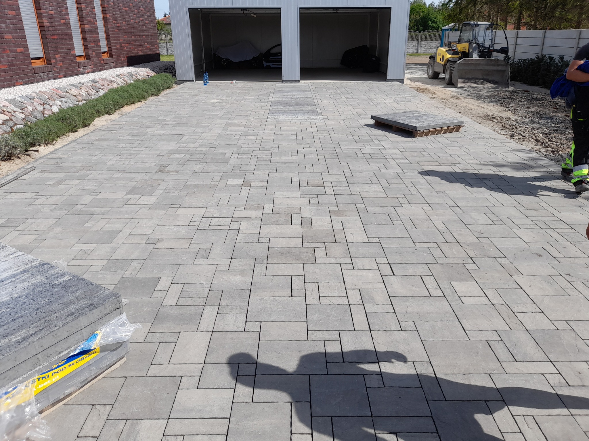Ułożona szara kostka brukowa o nieregularnym wzorze na podjeździe, z zaparkowanymi samochodami w garażu i miniładowarką w tle