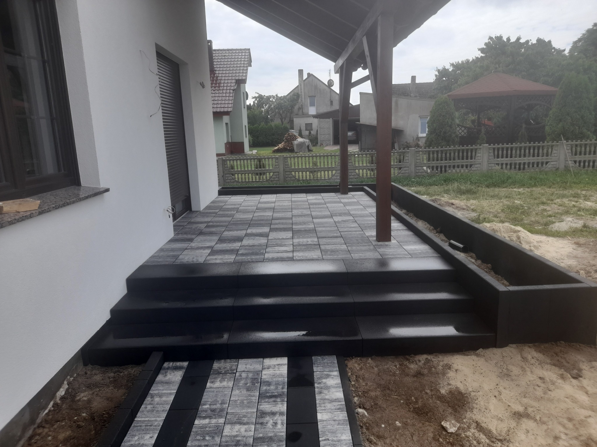 Zadaszony taras z kostki brukowej w odcieniach szarości i czerni, schody prowadzące do domu, widok na ogród z altaną i drewnem na opał.