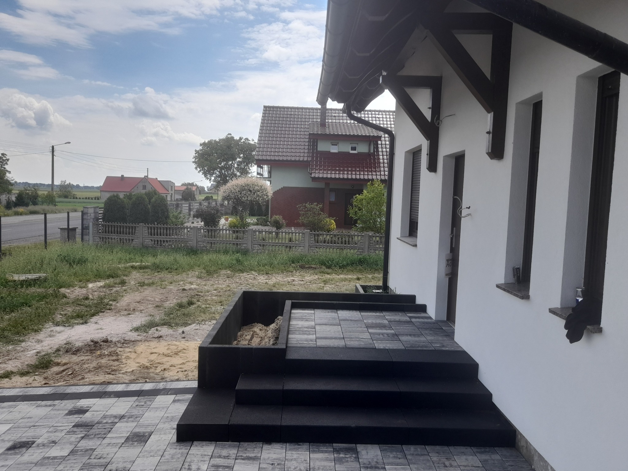 Nowoczesne, grafitowe schody wejściowe z kostki brukowej prowadzące do białego domu z elementami drewnianymi, widok z boku na tle zabudowy jednorodzinnej i błękitnego nieba.