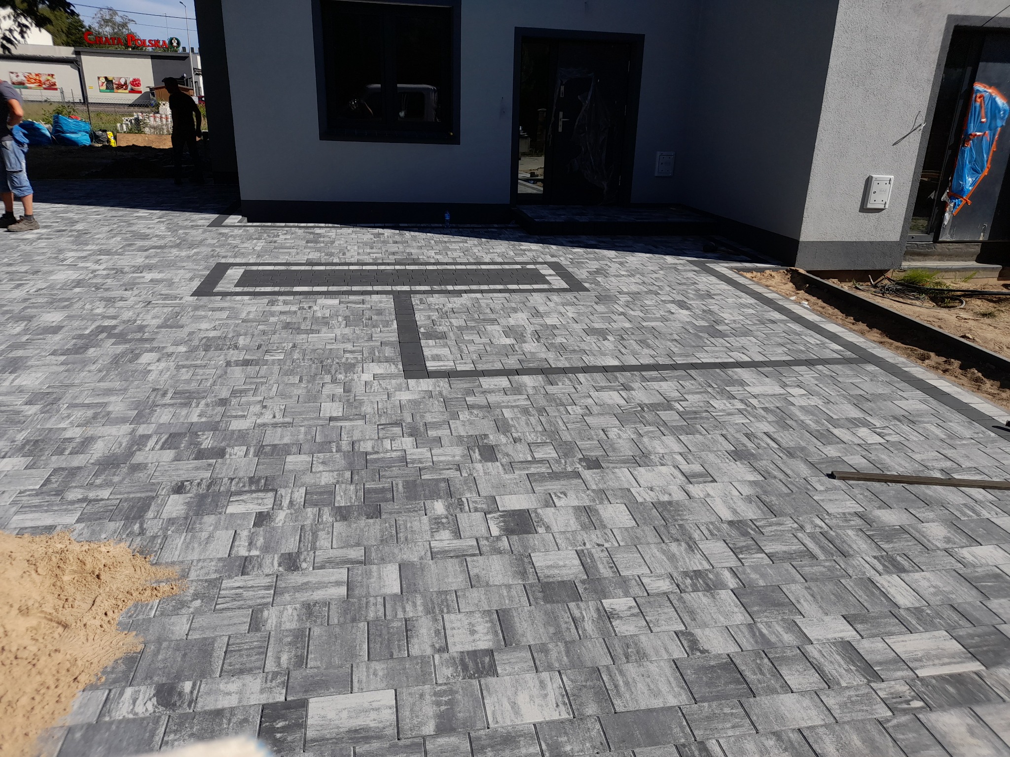 Ułożona szara kostka brukowa z ciemniejszymi obramowaniami przed budynkiem, widoczny fragment piasku i pracownicy w tle.