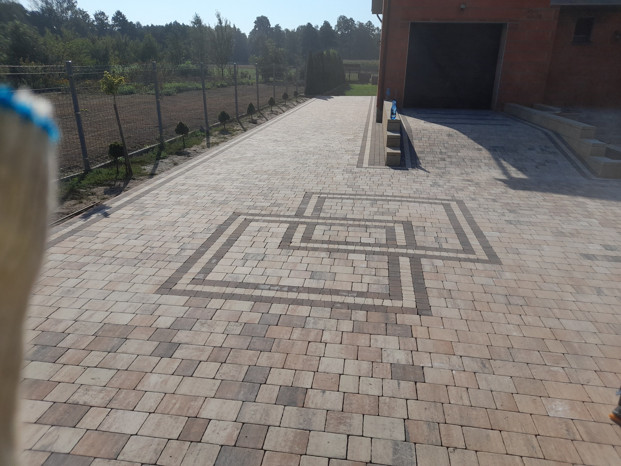 Ułożona kostka brukowa w odcieniach beżu i brązu tworząca geometryczny wzór przed garażem i ogrodzeniem.