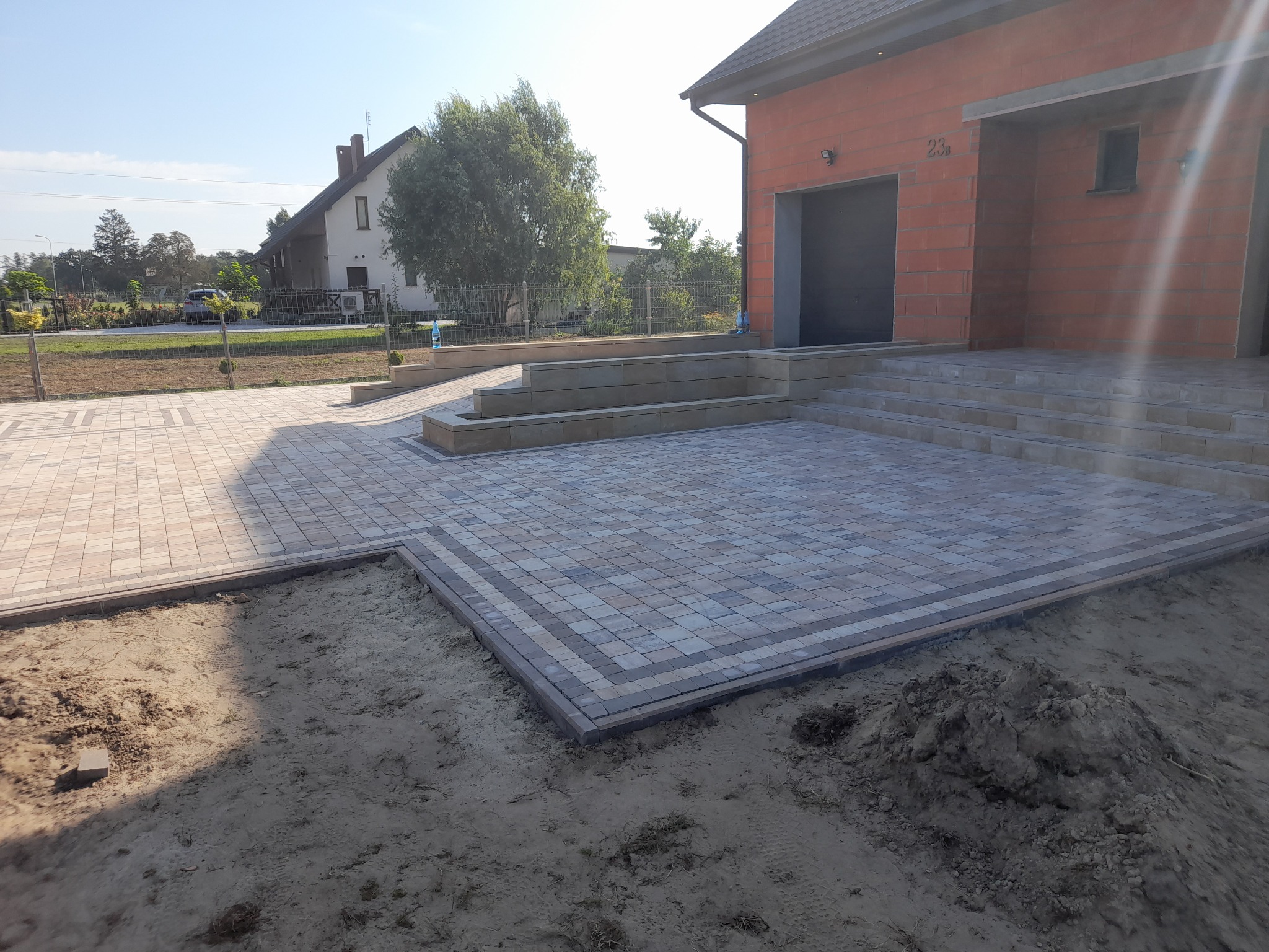 Ułożona kostka brukowa w neutralnych odcieniach przed nowoczesnym domem z garażem, widoczne schody wejściowe i przygotowany teren pod trawnik.