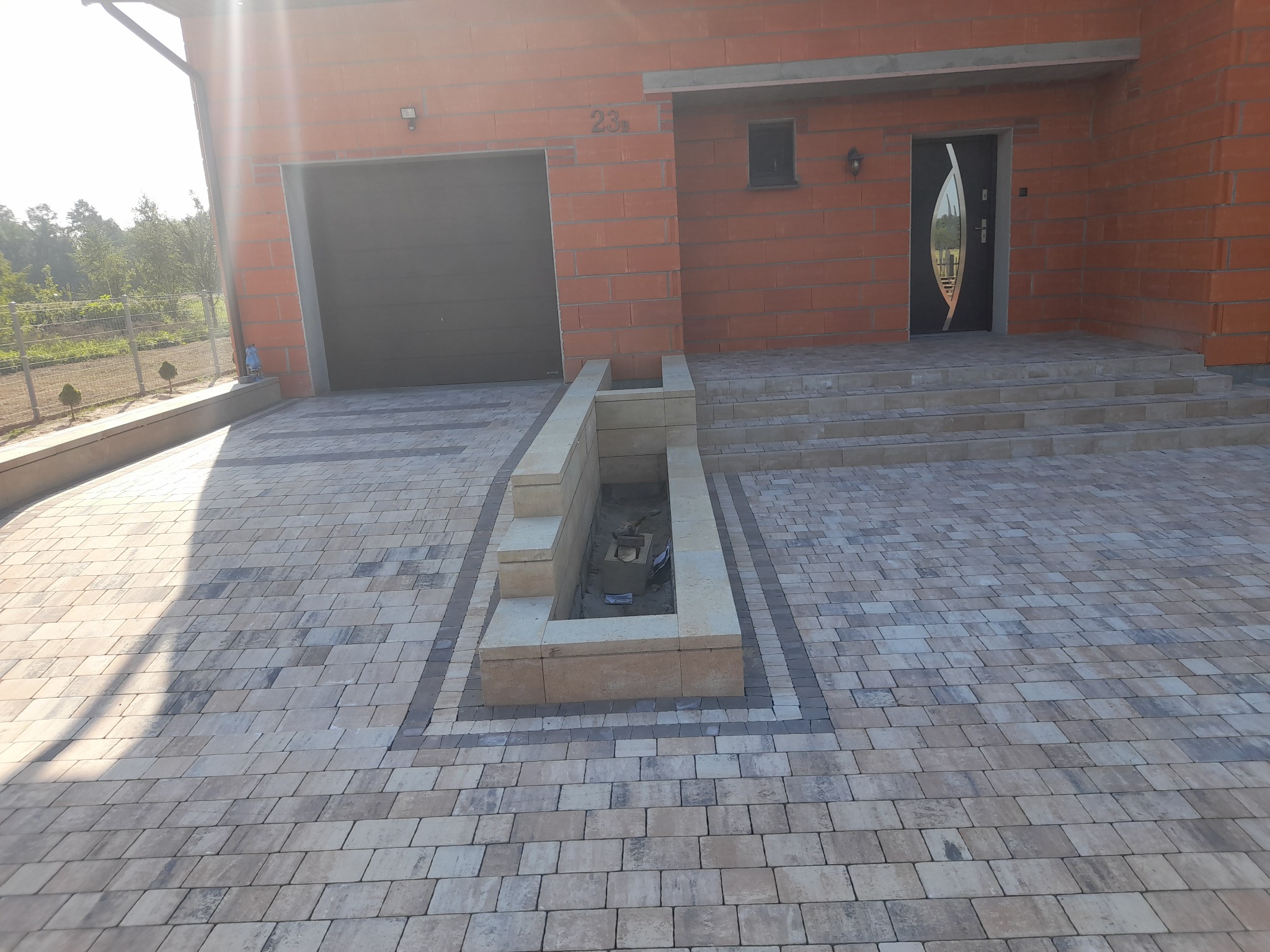 Ułożona kostka brukowa w odcieniach szarości i beżu prowadząca do domu z garażem, z elementami dekoracyjnymi w postaci obramowania z ciemniejszej kostki i podwyższonej rabaty.