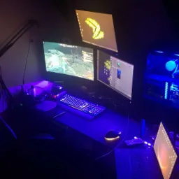 Stanowisko gracza z trzema monitorami, podświetlaną klawiaturą i obudową komputera z niebieskim oświetleniem LED, widok z góry, ciemne otoczenie.