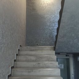 Sebastian Jasiński Budownictwo - Klatka schodowa z szarymi ścianami o dekoracyjnej fakturze, widok od dołu, oświetlenie sufitowe i przeszklone drzwi w tle.