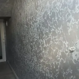 Ściana w pokoju pokryta dekoracyjną, srebrzystą farbą o nieregularnym wzorze, przypominającym popękany lód, widoczna z perspektywy narożnika pomieszczenia, z fragmentem podłogi i listwy...