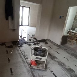 Sebastian Jasiński Budownictwo - Przygotowanie podłoża pod posadzkę w Wołuszewie: widoczna wylewka samopoziomująca na folii z logo producenta, narzędzia i pracownik w tle.