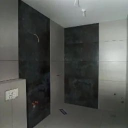 Wykończona łazienka z szarymi i ciemnymi płytkami imitującymi kamień, widoczne podłączenia hydrauliczne, odpływ liniowy w podłodze i biała stolarka okienna.