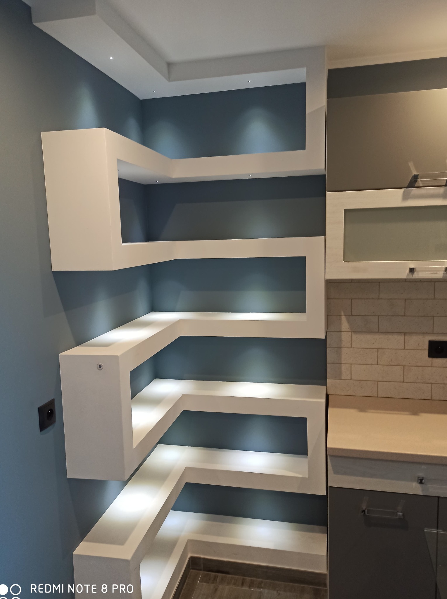 Narożna, podświetlana półka z płyty karton gips w nowoczesnej kuchni, zintegrowana z zabudową meblową i oświetleniem punktowym.