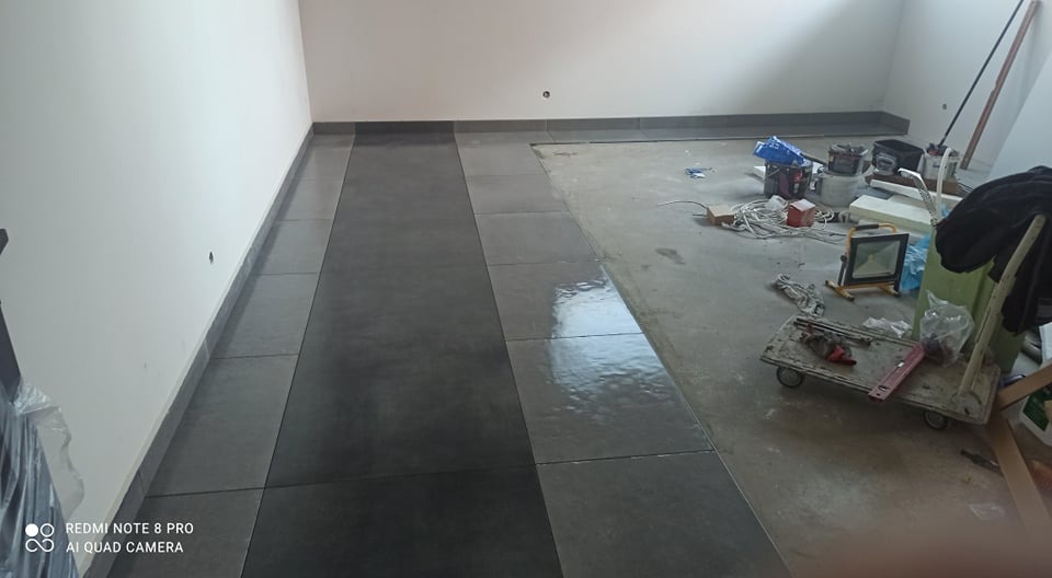 Układanie ciemnoszarych płytek podłogowych w pomieszczeniu, widoczny fragment ułożonej podłogi oraz przygotowane podłoże z narzędziami i materiałami budowlanymi.