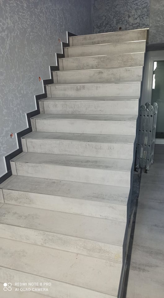 Schody wyłożone szarymi płytkami imitującymi beton, z czarnymi listwami przyściennymi i dekoracyjną szarą ścianą w tle, widoczna drabina aluminiowa oparta o ścianę.