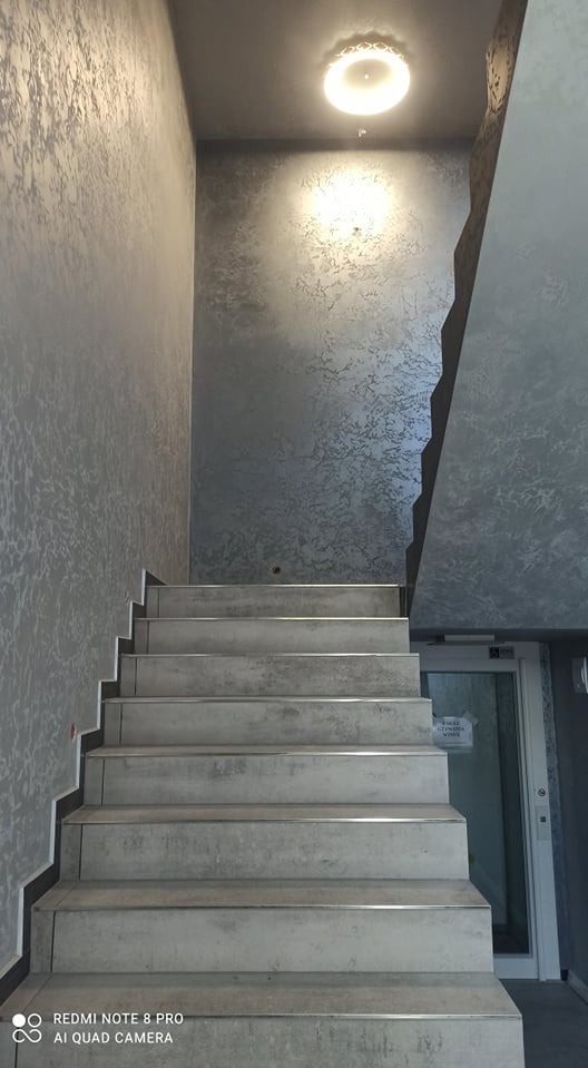 Klatka schodowa z szarymi ścianami o dekoracyjnej fakturze, widok od dołu, oświetlenie sufitowe i przeszklone drzwi w tle.