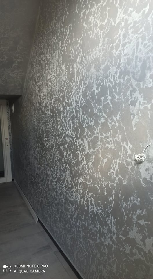 Ściana w pokoju pokryta dekoracyjną, srebrzystą farbą o nieregularnym wzorze, przypominającym popękany lód, widoczna z perspektywy narożnika pomieszczenia, z fragmentem podłogi i listwy...