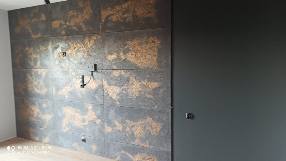 Dekoracyjna ściana z paneli imitujących beton z nieregularnymi złotymi wzorami, widoczne gniazdka elektryczne i wyprowadzone kable, obok ściana pomalowana na jednolity szary kolor.