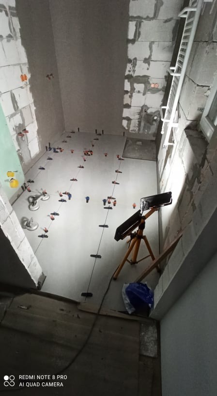 Układanie jasnych płytek podłogowych w surowym wnętrzu, widoczne narzędzia glazurnicze, system poziomowania płytek, lampa budowlana na trójnogu oświetla kładzioną podłogę.