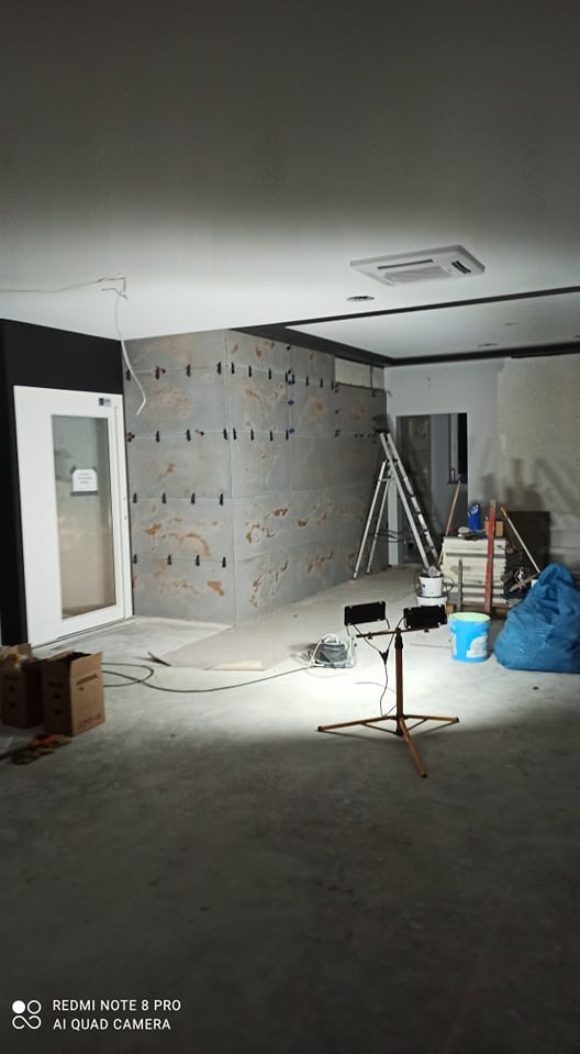 Montaż płyt imitujących kamień na ścianie z widocznymi uchwytami montażowymi, drabina, lampa budowlana i akcesoria malarskie w pomieszczeniu w trakcie remontu.