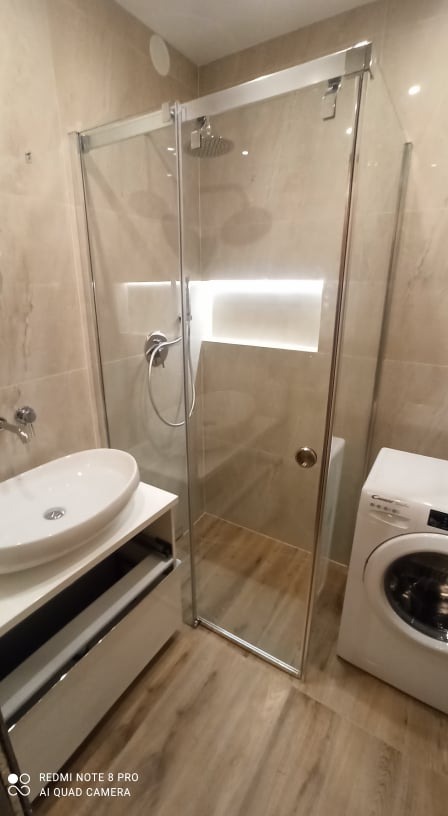 Nowoczesna łazienka z kabiną prysznicową typu walk-in, umywalką nablatową na szafce z szufladami i pralką, wykończona płytkami w odcieniach beżu i brązu z drewnopodobną podłogą.