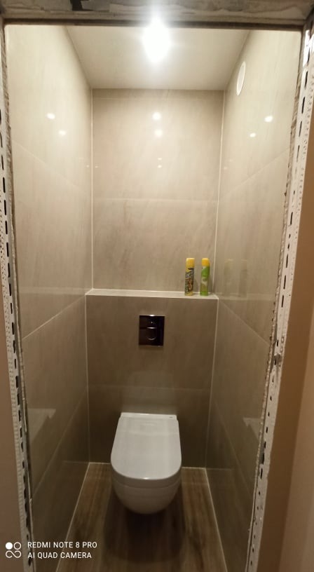 Wykończona toaleta w neutralnych barwach z podwieszaną muszlą klozetową, płytkami imitującymi kamień i metalowymi listwami wykończeniowymi przy ościeżnicy.