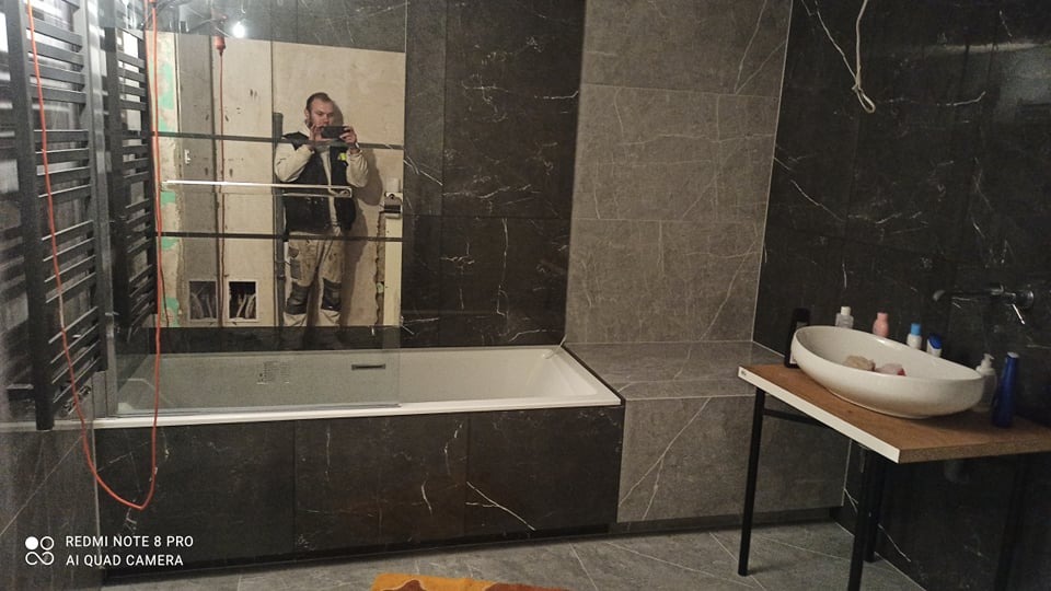 Wnętrze łazienki z ciemnymi marmurkowymi płytkami, białą wanną z parawanem i owalną umywalką na drewnianym stoliku, widok w lustrze na osobę robiącą zdjęcie podczas prac wykończeniowych.