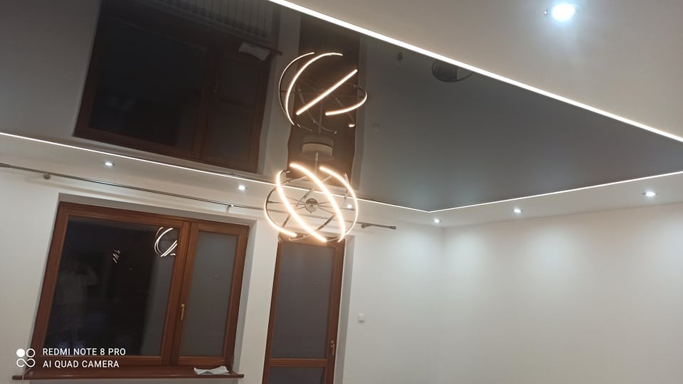 Błyszczący, ciemny sufit napinany z oświetleniem LED liniowym i kulistą lampą wiszącą z metalowymi obręczami, odbijający okna w pokoju.