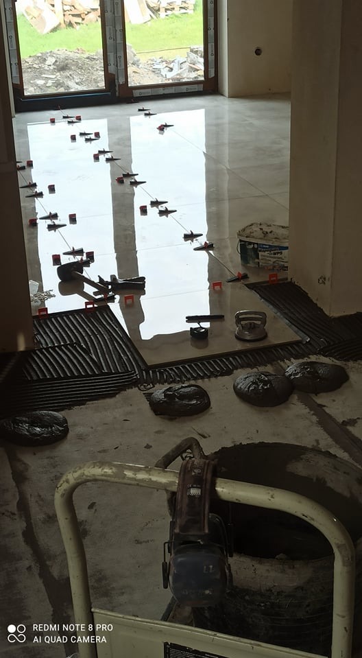 Układanie błyszczących płytek podłogowych w jasnym pomieszczeniu, widoczne narzędzia, poziomice i krzyżyki dystansowe, w tle okno z widokiem na zieleń.