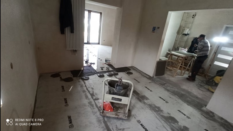 Przygotowanie podłoża pod posadzkę w Wołuszewie: widoczna wylewka samopoziomująca na folii z logo producenta, narzędzia i pracownik w tle.