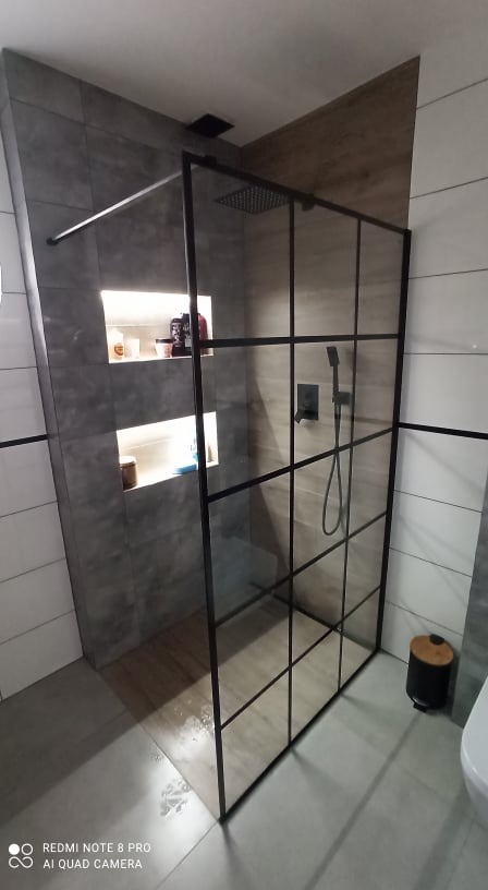 Nowoczesna kabina prysznicowa z czarnymi profilami i deszczownicą, wbudowane półki w szarej płytce, widok na baterię i słuchawkę prysznicową, jasne płytki podłogowe.