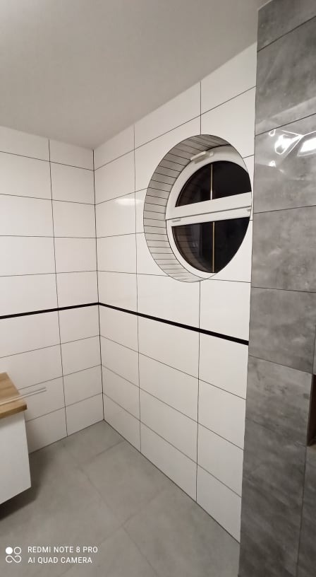 Wykończona łazienka z białymi, prostokątnymi płytkami, okrągłym oknem i szarym panelem na ścianie