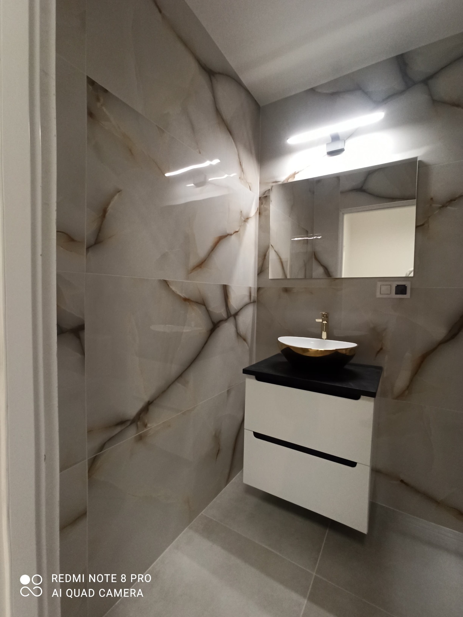 Wykończona łazienka z marmurowymi płytkami na ścianach, białą szafką z czarnym blatem i owalną umywalką w kolorze białym ze złotym wykończeniem, widok na lustro z oświetleniem.