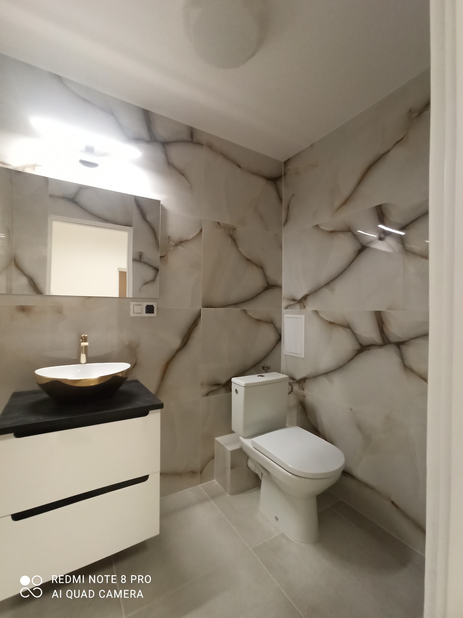 Wykończona łazienka z marmurowymi płytkami, złotą umywalką na czarnym blacie i białą szafką, widok na toaletę i lustro.