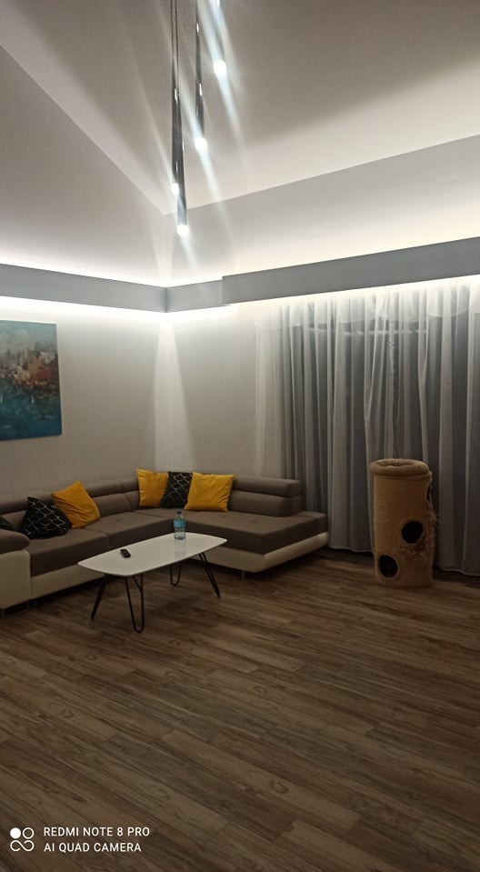 Nowoczesny salon z narożną sofą, żółtymi poduszkami i drewnianą podłogą; oświetlenie sufitowe LED i abstrakcyjny obraz na ścianie.