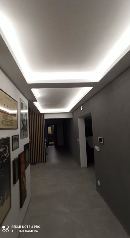 Nowoczesny korytarz z podwieszanym sufitem i oświetleniem LED, szara ściana, obrazy na ścianie po lewej stronie, widok w głąb mieszkania przez drewniane lamele.