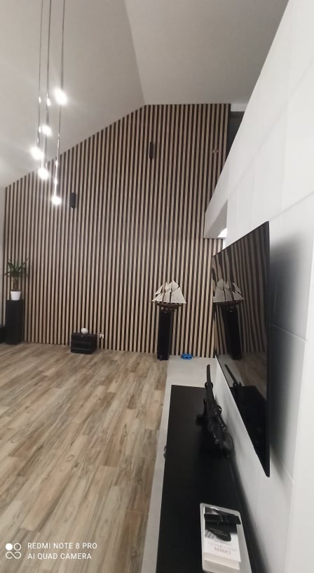 Nowoczesny salon z drewnianą ścianą z paneli pionowych, drewnianą podłogą i czarnymi akcentami dekoracyjnymi, takimi jak model żaglowca i wysoki stojak na rośliny.