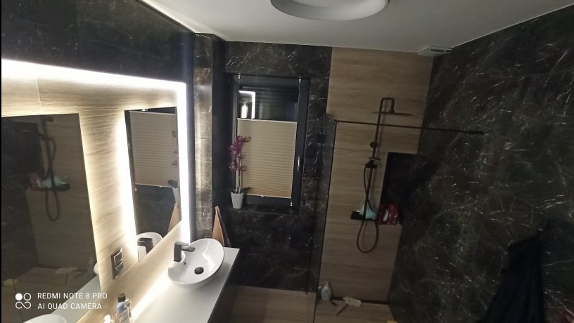 Nowoczesna łazienka z czarno-beżowymi marmurowymi płytkami, podświetlanym lustrem nad białą umywalką nablatową, kabiną prysznicową typu walk-in z czarną armaturą i storą w oknie.