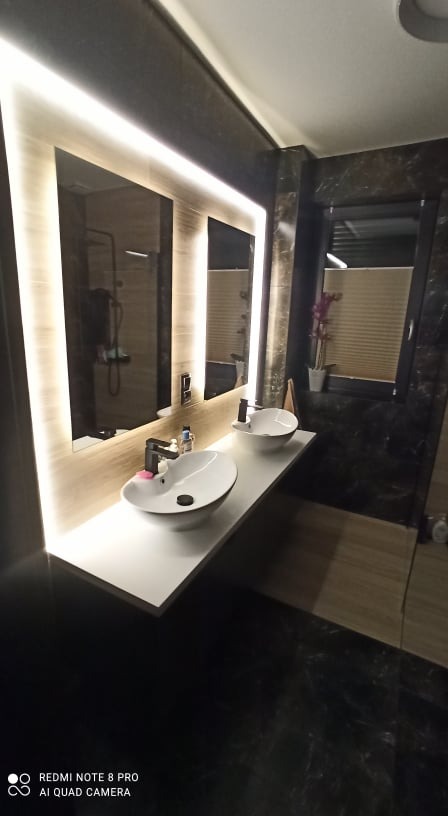 Nowoczesna łazienka z dwoma owalnymi umywalkami na białym blacie, podświetlane lustra i marmurowe wykończenie ścian oraz podłogi.