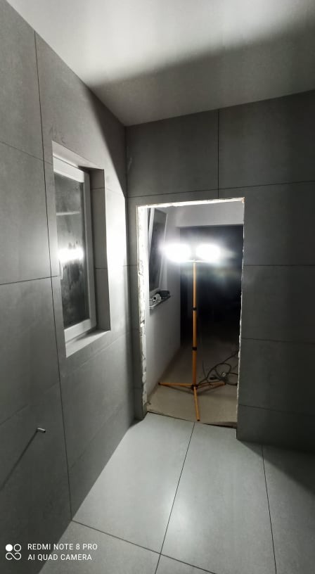 Wnętrze w trakcie wykańczania płytkami w odcieniach szarości, widoczna lampa budowlana oświetlająca przejście i fragment okna.
