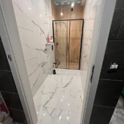 Wnętrze łazienki w trakcie remontu: widoczne białe płytki imitujące marmur z brązowymi żyłami, kabina prysznicowa z czarnymi ramami, sufit w trakcie szpachlowania, niedokończone obramowanie drzwi.