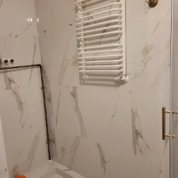 Biała łazienka wykończona płytkami imitującymi marmur, z grzejnikiem drabinkowym, miedzianymi rurami i pomarańczową gąbką na brodziku.