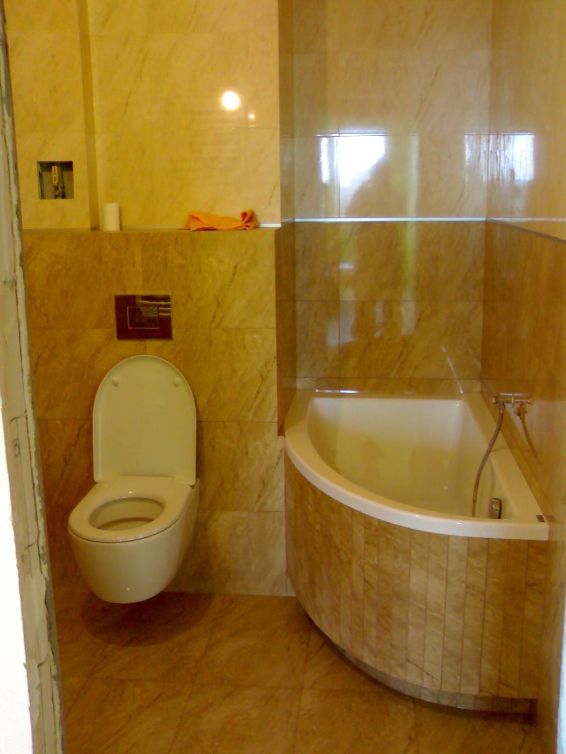 Wykończona łazienka w beżowych płytkach imitujących marmur, z wiszącą toaletą i narożną wanną obudowaną płytkami, pomarańczowy ręcznik na półce.