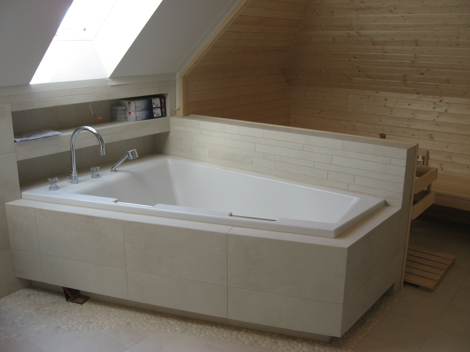 Wanna obudowana beżowymi płytkami w łazience na poddaszu z drewnianą boazerią i oknem dachowym, widoczna armatura i półka z akcesoriami.