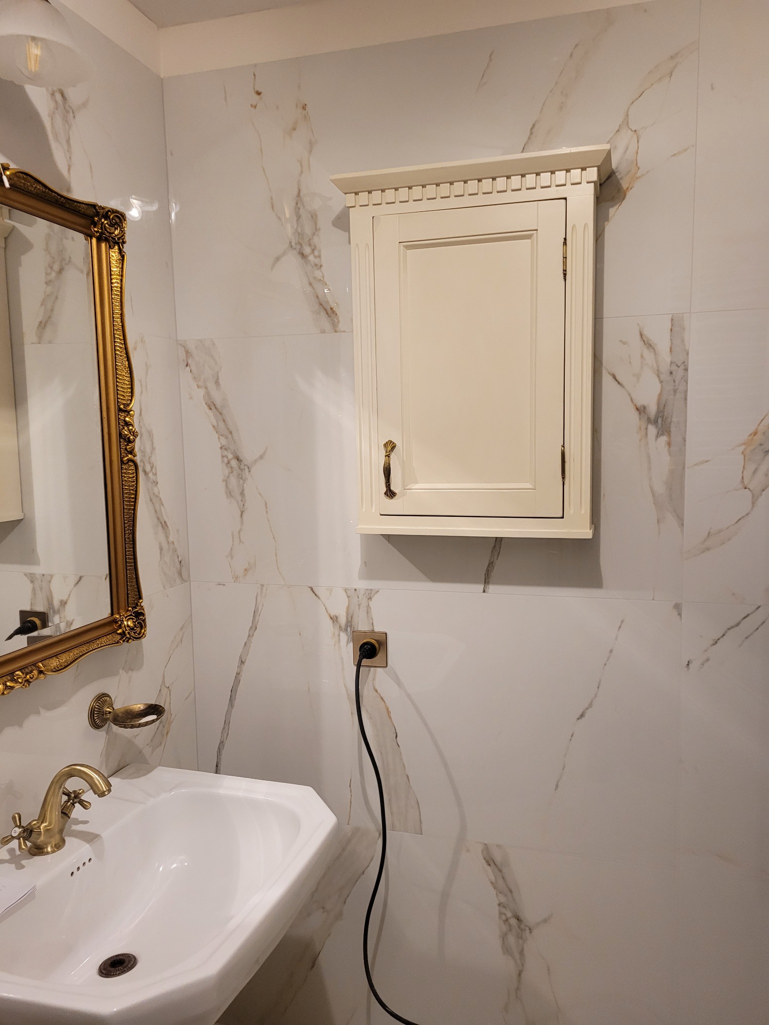 Łazienka z białymi marmurkowymi płytkami, kremową szafką nad umywalką i złotą armaturą oraz ozdobnym lustrem.