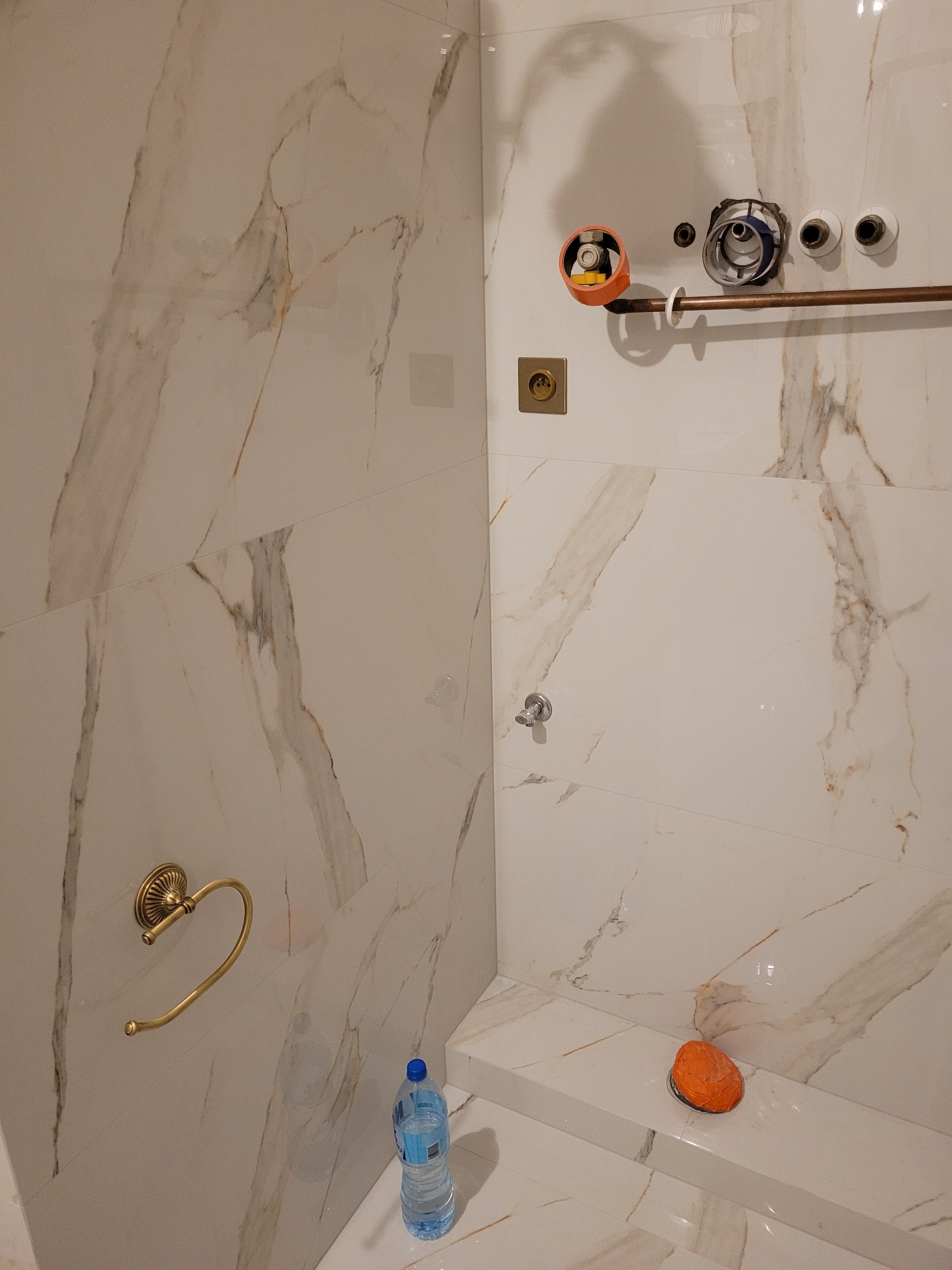 Wnętrze łazienki w trakcie prac wykończeniowych, widoczne białe płytki imitujące marmur, zamontowany uchwyt na ręcznik w kolorze złotym, przygotowane przyłącza wodne i butelka wody mineralnej...