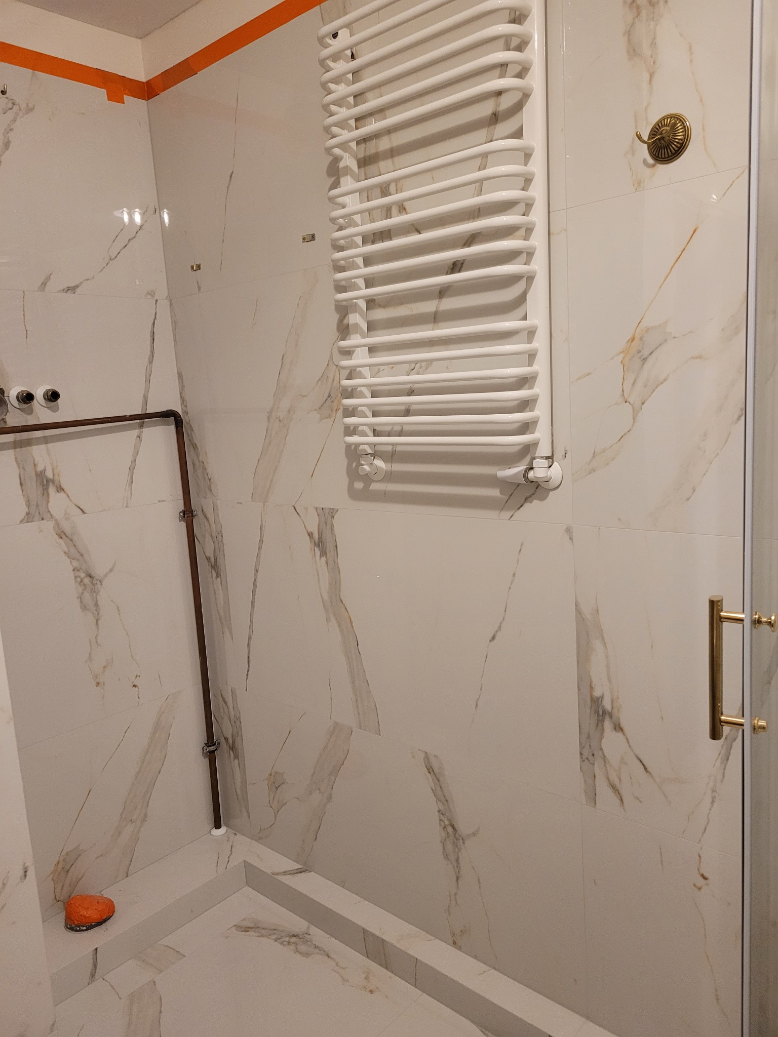 Biała łazienka wykończona płytkami imitującymi marmur, z grzejnikiem drabinkowym, miedzianymi rurami i pomarańczową gąbką na brodziku.