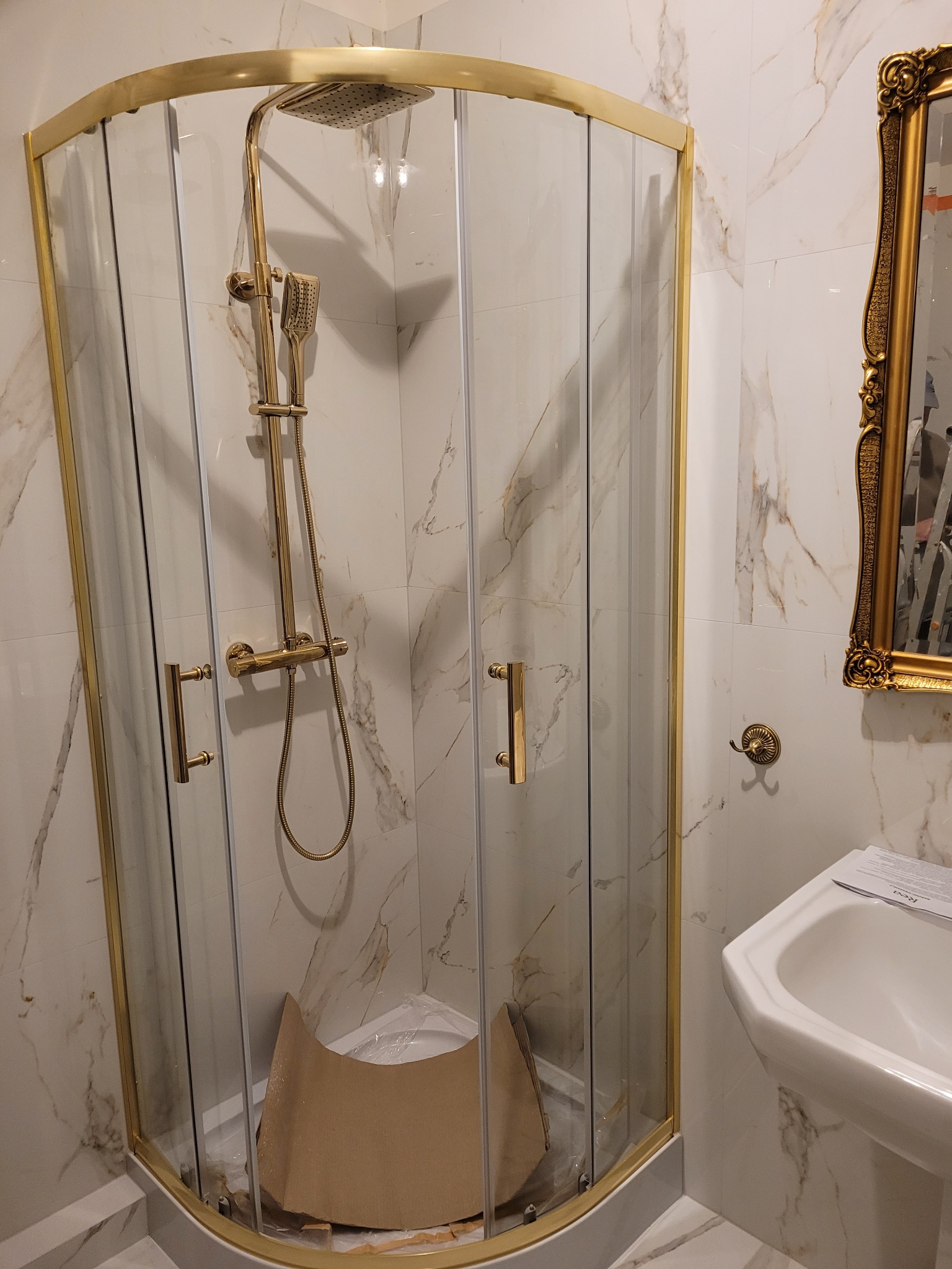 Złota kabina prysznicowa narożna z deszczownicą i baterią, marmurowe płytki na ścianach, widoczna umywalka i złote lustro w ozdobnej ramie, ochrona brodzika z kartonu.