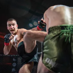 Zawodnik MMA w trakcie kopnięcia, z ochraniaczem na zęby i tatuażem na ramieniu, widoczny przeciwnik w zielonych spodenkach.