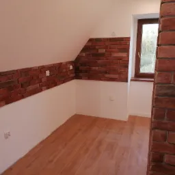 Pokój na poddaszu z wykończoną ścianą z cegły klinkierowej, drewnianą podłogą i oknem z widokiem na drzewa.