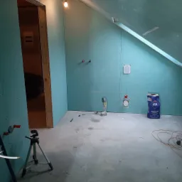 Wnętrze łazienki w trakcie montażu płyt gipsowo-kartonowych na ścianach, widoczne wyprowadzenia instalacji wodnej, laser krzyżowy na statywie, oraz worek zaprawy klejowej Atlas