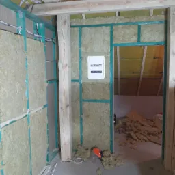 Wnętrze w trakcie prac: ściany z izolacją z wełny mineralnej, widoczne drewniane belki konstrukcyjne, instalacja elektryczna w puszce oraz rozrzucona wełna mineralna na podłodze i narzędzie...