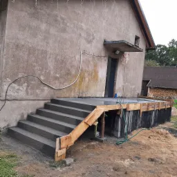 Surowe betonowe schody prowadzące do drewnianego tarasu przy szarej elewacji budynku gospodarczego, widoczne drewniane szalunki i czarna izolacja fundamentów.