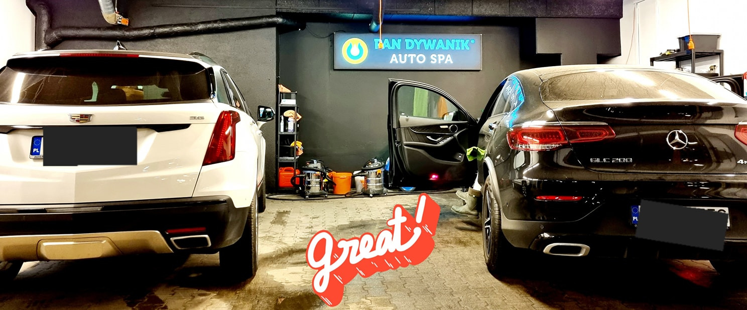 Dwa samochody, biały Cadillac XT5 i czarny Mercedes GLC 200, stoją w myjni samochodowej, z otwartymi drzwiami w Mercedesie, widoczne urządzenia do czyszczenia i napis 'Pan Dywanik Auto Spa' w tle.